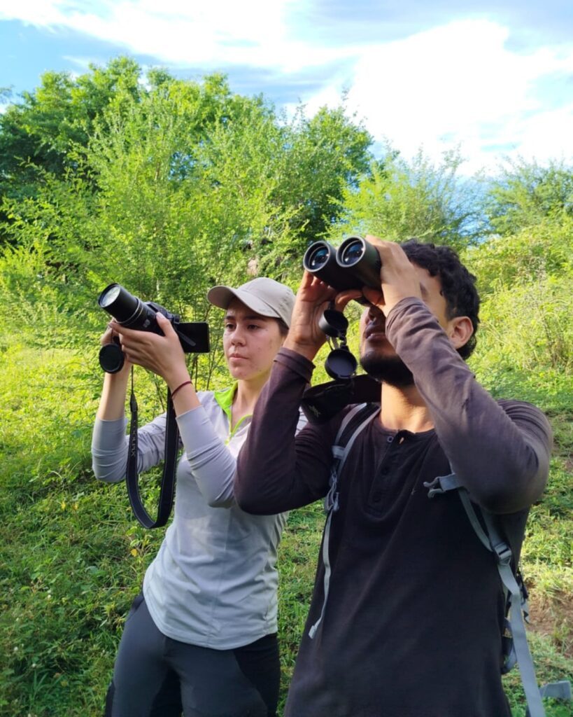 Dos participantes observan aves con binoculares y cámara en una zona boscosa